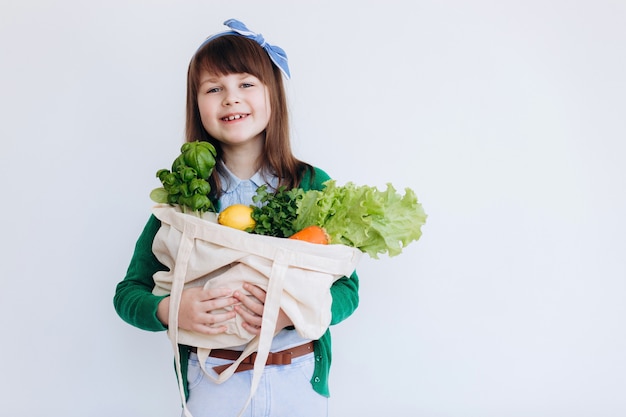 Niña sosteniendo una bolsa de supermercado textil con verduras. Concepto de desperdicio cero. Compra de alimentos sin empaque. Bolsa natural ecológica con frutas y verduras orgánicas.