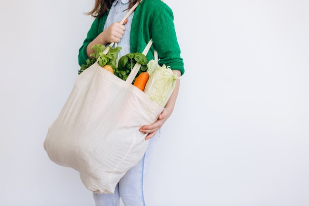 Niña sosteniendo una bolsa de supermercado textil con verduras. Concepto de desperdicio cero. Compra de alimentos sin empaque. Bolsa natural ecológica con frutas y verduras orgánicas.