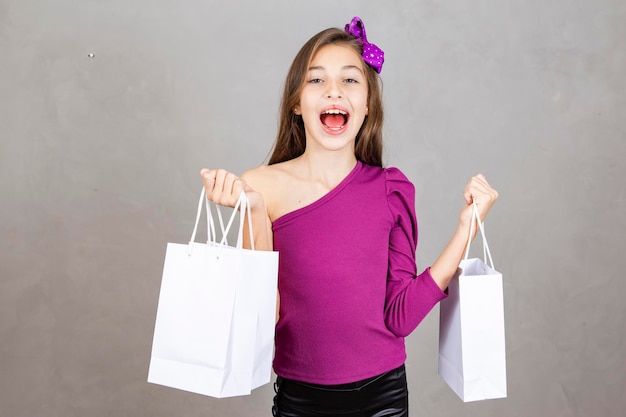 Niña sosteniendo bolsas de compras de pie aislado sobre fondo gris Niña de unos diez años con bolsas de compras