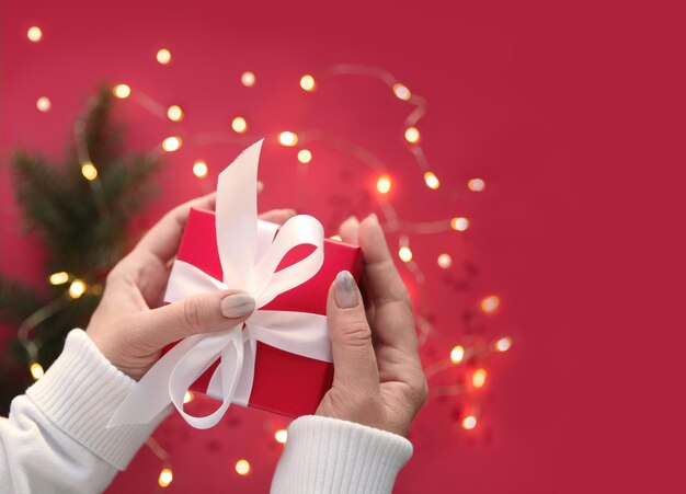 Una niña sosteniendo una caja de regalo roja decorada con una cinta blanca en el fondo de una guirnalda borrosa