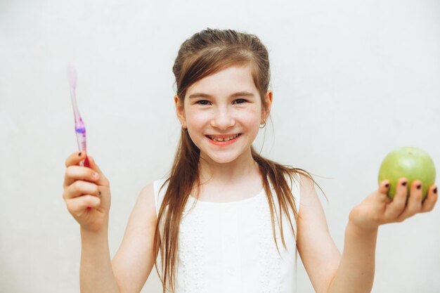 Niña sosteniendo un cepillo de dientes y una manzana sobre un fondo blanco El concepto de cepillarse los dientes La cara sonriente de una niña sostiene una manzana y un cepillo de dientes la cara feliz del niño se encarga de la higiene