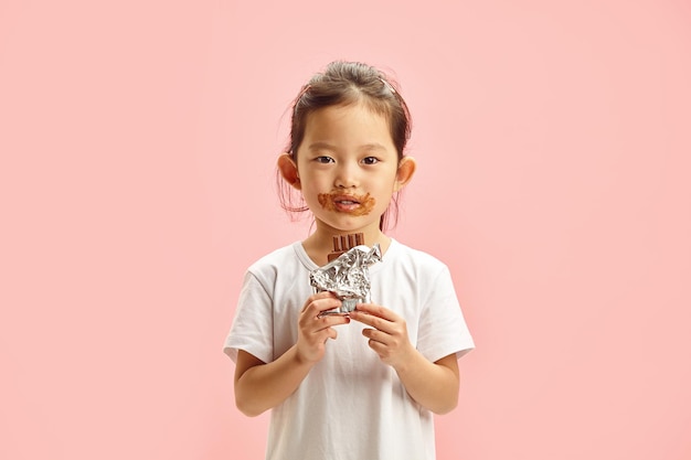 Foto niña sosteniendo y comiendo chocolate en baldosas manchado alrededor de su boca de pie sobre rosa aislado
