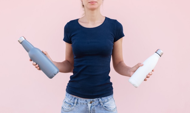 Foto niña sosteniendo dos botellas de agua termo de acero reutilizables, blanco y gris de colores. fondo rosa pastel. sea libre de plástico. cero desperdicio.