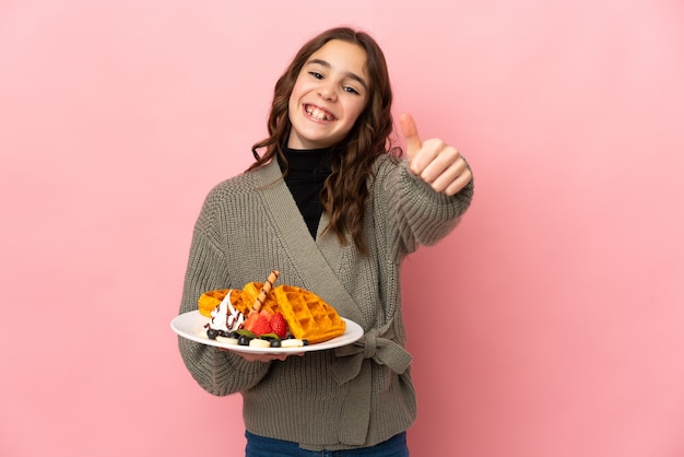 Foto niña sosteniendo gofres aislado sobre fondo rosa con los pulgares hacia arriba porque ha sucedido algo bueno