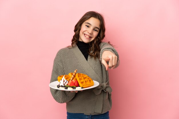 Niña sosteniendo gofres aislados sobre fondo rosa apuntando al frente con expresión feliz