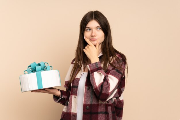 Niña sosteniendo un gran pastel aislado en beige con dudas