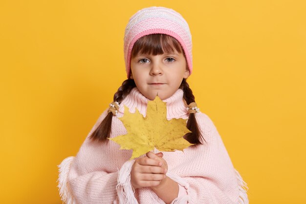 Niña sosteniendo hojas de otoño y mirando directamente al frente, vistiendo poncho y gorra rosa