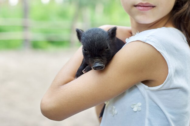 Niña sosteniendo en manos de lechón bebé recién nacido negro. Granja, agricultura, país, mascotas, concepto de niños