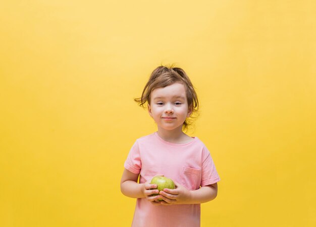 Una niña sosteniendo una manzana verde en un espacio amarillo. Linda chica con una cola de caballo en una camiseta rosa. Buena nutricion. Copia espacio