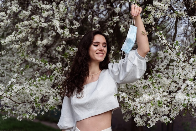 Niña sosteniendo una máscara médica después del final del período de cuarentena por coronavirus covid-19. Modelo posando cerca de árboles florecientes blancos en el jardín.