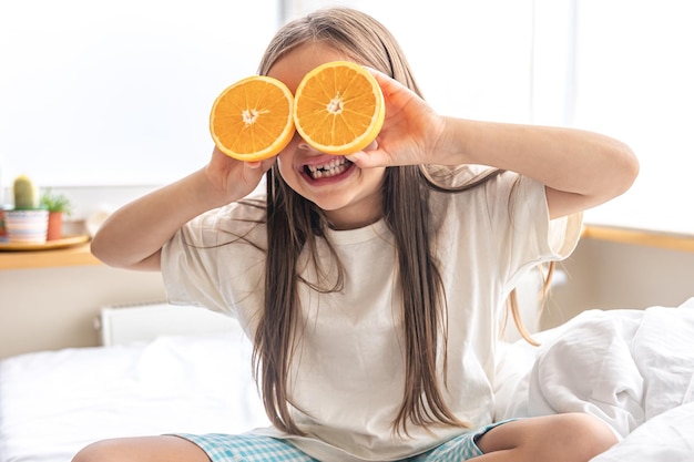 Niña sosteniendo naranjas cerca de sus ojos mientras está sentada en la cama