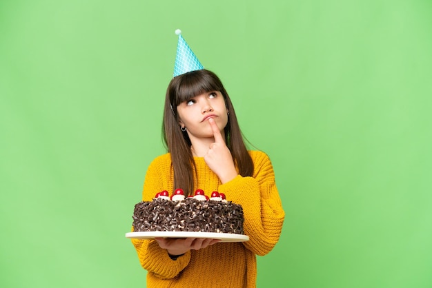 Niña sosteniendo pastel de cumpleaños sobre un fondo aislado de clave de croma que tiene dudas mientras mira hacia arriba