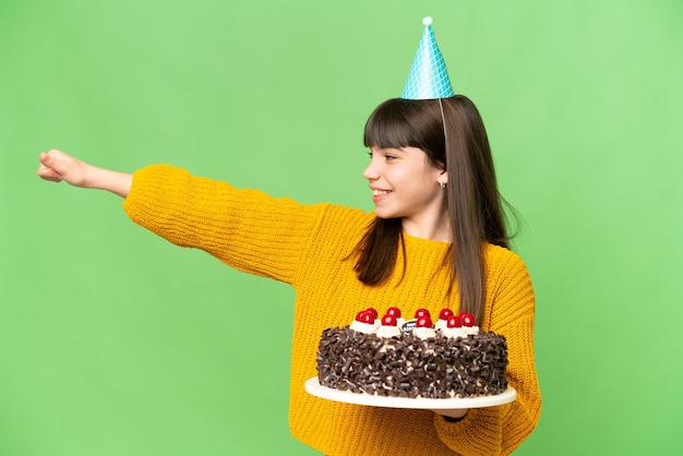Niña sosteniendo pastel de cumpleaños sobre un fondo de clave de croma aislado dando un gesto de pulgar hacia arriba