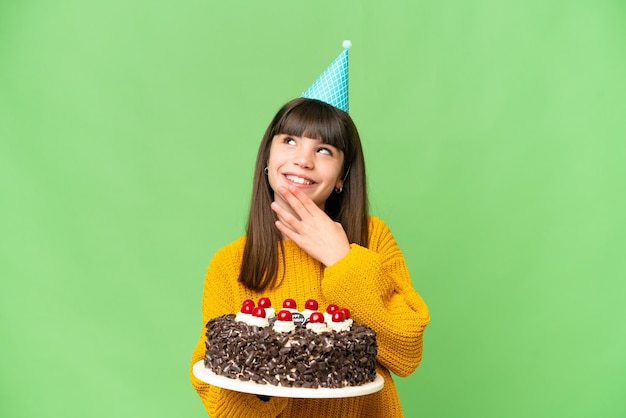 Niña sosteniendo pastel de cumpleaños sobre un fondo de clave de croma aislado mirando hacia arriba mientras sonríe