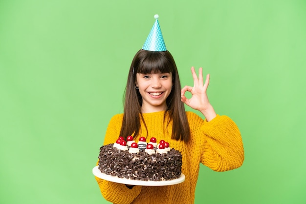Niña sosteniendo pastel de cumpleaños sobre fondo de clave de croma aislado que muestra el signo ok con los dedos