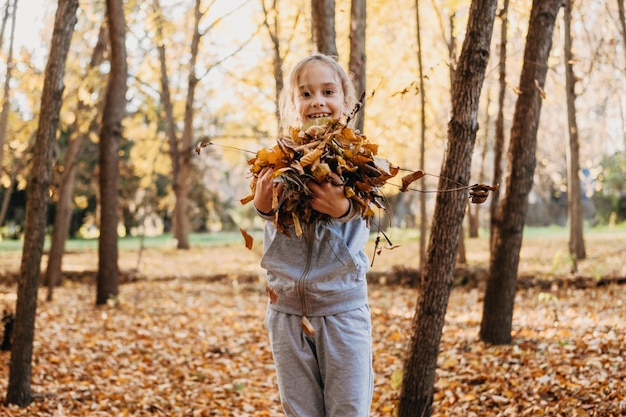 Foto niña sosteniendo un pequeño montón de hojas amarillas caídas en el bosque de otoño sonriendo mirando a la cámara montón de hojas en el césped los niños ayudan con las tareas los niños juegan al aire libre