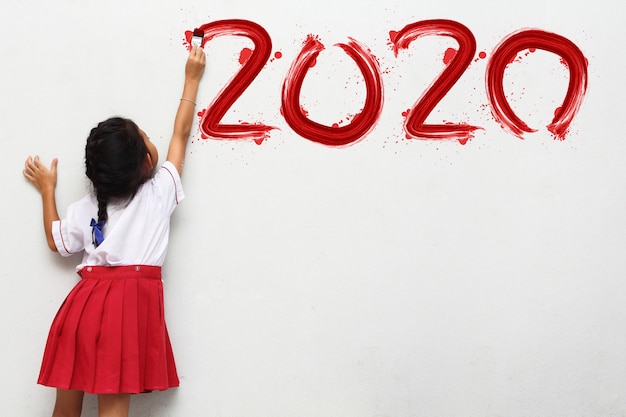 Niña sosteniendo un pincel pintando feliz año nuevo 2020 en una pared blanca