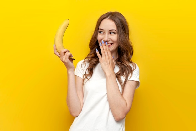 Foto niña sosteniendo un plátano pequeño y burlándose de un fondo amarillo aislado mujer presenta erótica