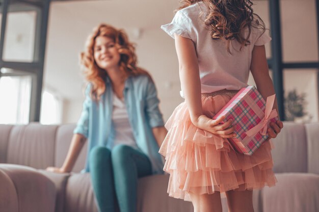 Niña sosteniendo un regalo para mamá sentada en el sofá detrás de la espalda en casa