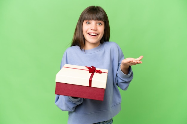 Niña sosteniendo un regalo sobre un fondo clave de croma aislado con una expresión facial sorprendida