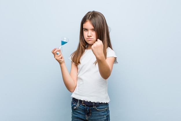 Niña sosteniendo un reloj de arena mostrando el puño a la cámara, expresión facial agresiva