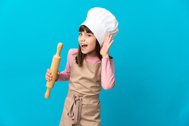 Foto niña sosteniendo un rodillo aislado en azul escuchando algo poniendo la mano en la oreja