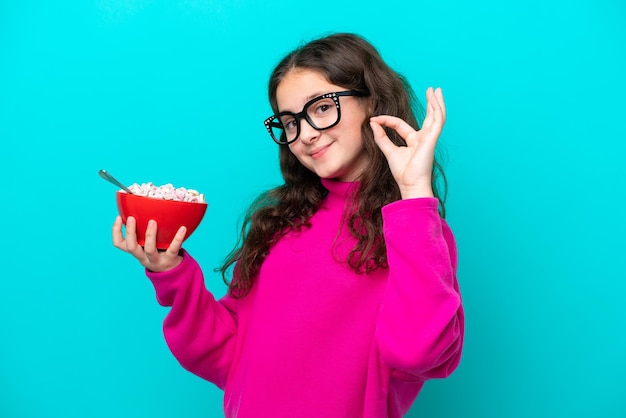 Niña sosteniendo un tazón de cereales aislado sobre fondo azul que muestra el signo de ok con los dedos