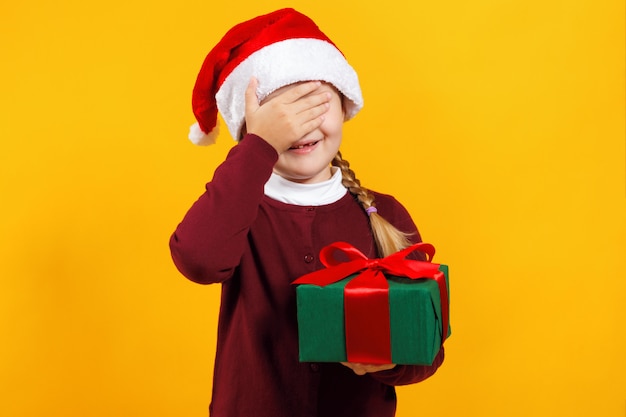 La niña sostiene una caja con un regalo y cerró los ojos con la mano.