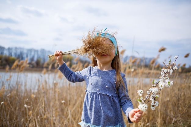 Niña sostiene cañas secas y una rama con pequeñas flores blancas en las manos, clima soleado de primavera, smilling y alegría del niño