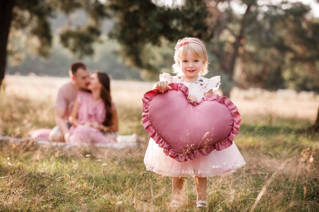 Niña sostiene un gran corazón rosa en el parque de verano mientras los padres se abrazan en el fondo