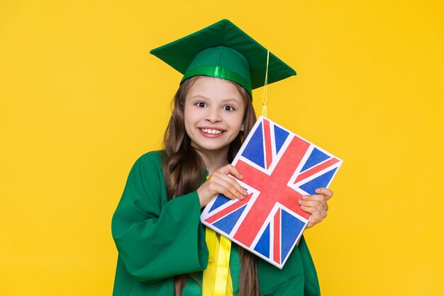 Foto una niña sostiene un libro en inglés con la bandera de gran bretaña un estudiante con sombrero de maestro se regocija en la graduación escuela de idiomas para niños fondo amarillo aislado
