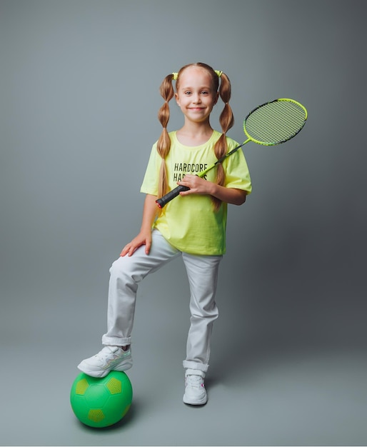 Una niña sostiene una pelota y una raqueta sobre un fondo gris. El concepto de deportes profesionales.