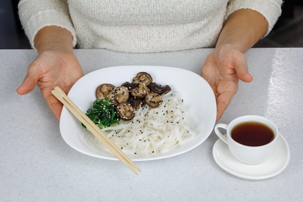 La niña sostiene un plato de fideos de arroz y setas shiitake en sus manos Taza blanca con café