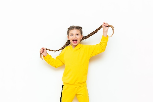 Una niña sostiene su cabello largo en trenzas en sus manos y sonríe ampliamente Un niño con un traje deportivo amarillo sobre un fondo blanco aislado
