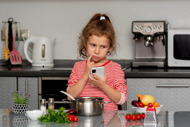 Una niña sostiene un teléfono y piensa qué cocinar con verduras frescas. Fotografía conceptual.