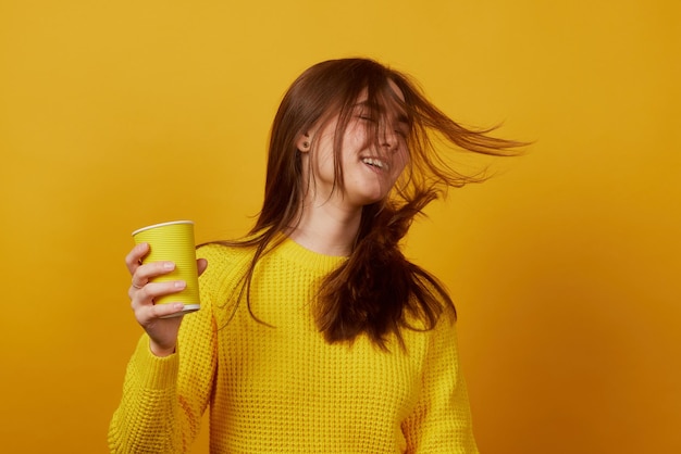 La niña sostiene un vaso desechable en la mano y se regocija con un fondo amarillo.