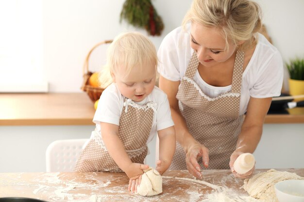 Niña y su madre rubia con delantales beige jugando y riendo mientras amasan la masa en la cocina. Pastelería casera para pan, pizza o galletas horneadas. Diversión familiar y concepto de cocina.