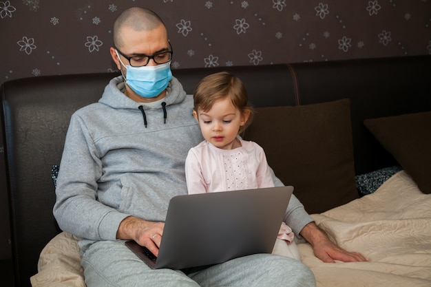 Niña y su padre en la máscara con el portátil en casa