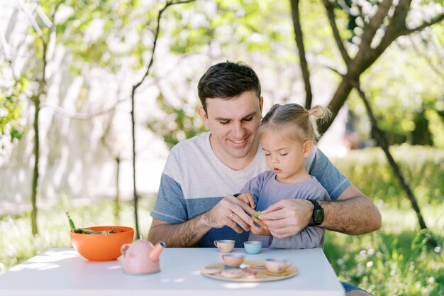 Foto niña con su padre pelando frijoles verdes mientras está sentada en una mesa en una fiesta de té en el jardín