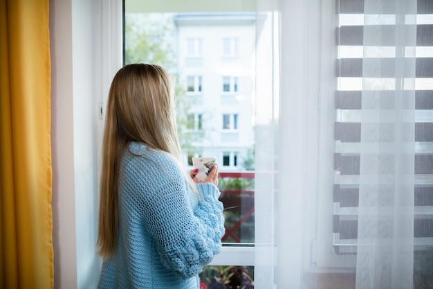 La niña se para con su taza junto a la ventana y mira afuera.