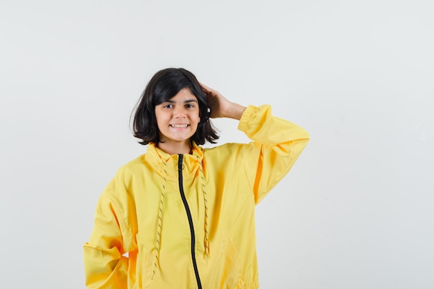 Niña en sudadera con capucha amarilla sosteniendo la mano en la cabeza y mirando alegre, vista frontal.