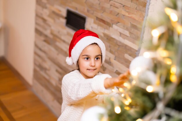 Niña en suéter adornos colgantes en el árbol de Navidad con luz, chuchería. Niño decorando el árbol de Navidad en la hermosa sala de estar familiar con chimenea.