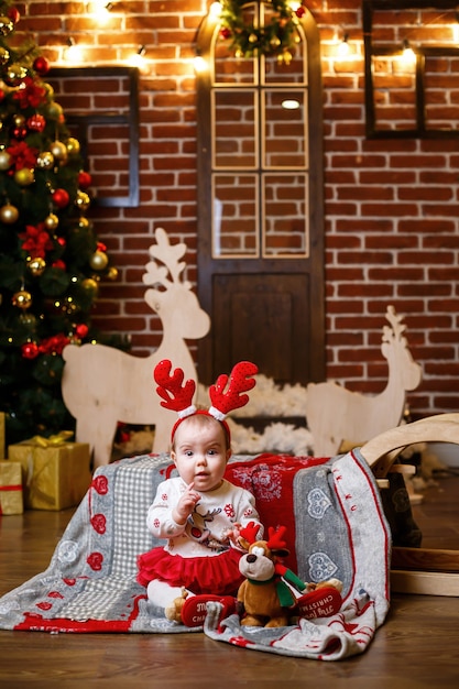 Una niña con un suéter cálido se sienta debajo de un árbol de Navidad con juguetes y regalos con cuernos en la cabeza. Infancia feliz. Ambiente de vacaciones de año nuevo