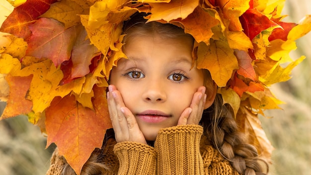 Una niña con un suéter y una corona de arce en la cabeza en otoño.