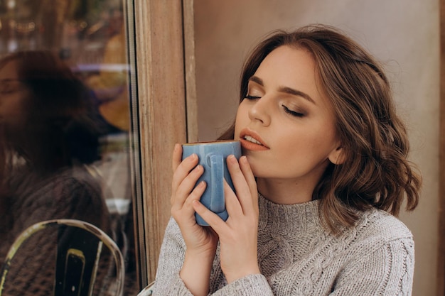 Una niña con un suéter gris se sienta en una mesa en una cafetería y bebe cacao y disfruta del aroma.