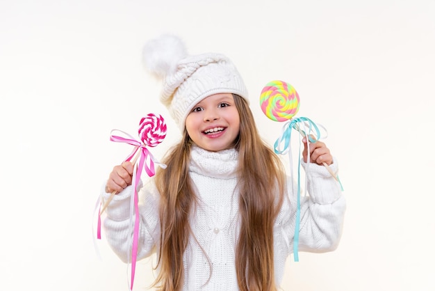 Una niña con un suéter de invierno sostiene una deliciosa piruleta y sonríe en un fondo blanco