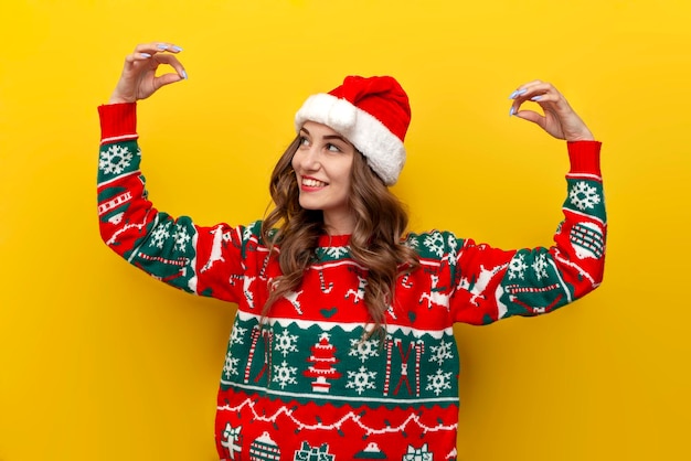 Niña con suéter de Navidad y sombrero de Papá Noel sostiene las manos vacías en un fondo aislado amarillo