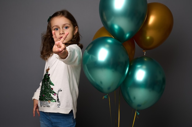 Niña con suéter navideño se encuentra cerca de hermosas bolas de aire doradas y metálicas de color verde brillante y muestra un signo de la paz, haciendo un gesto con el dedo, aislado sobre fondo gris con espacio de copia para anuncios