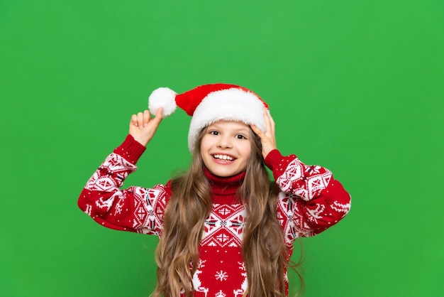 Una niña con un suéter navideño juega con un sombrero de Papá Noel antes de las vacaciones en un fondo verde aislado