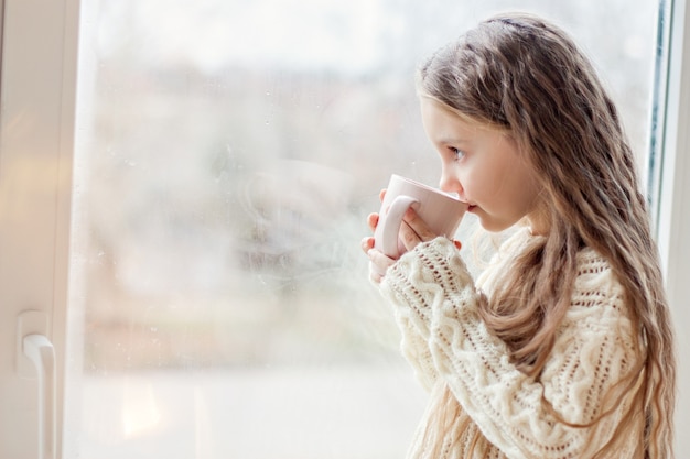 Una niña con un suéter de punto blanco bebe chocolate caliente, cacao